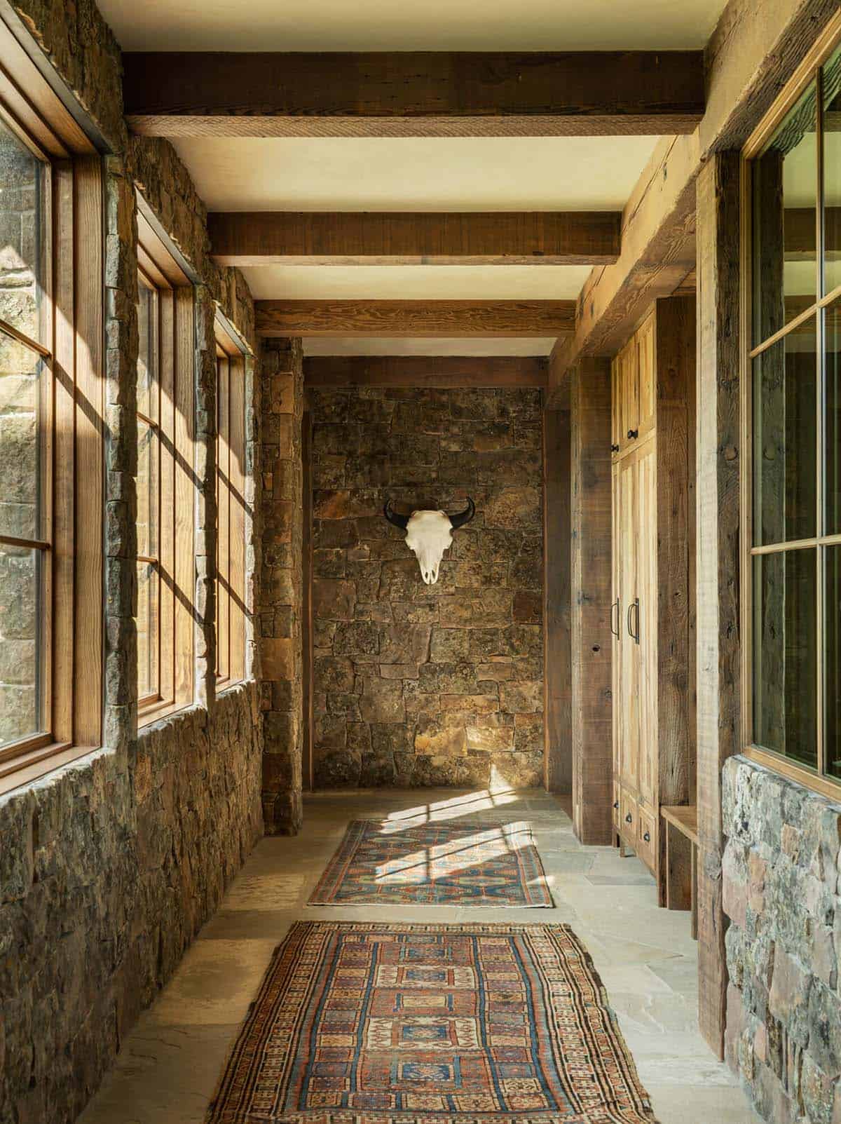 Stone-walled hallway with wood beams, layered patterned rugs, and a mounted cattle skull on the end wall