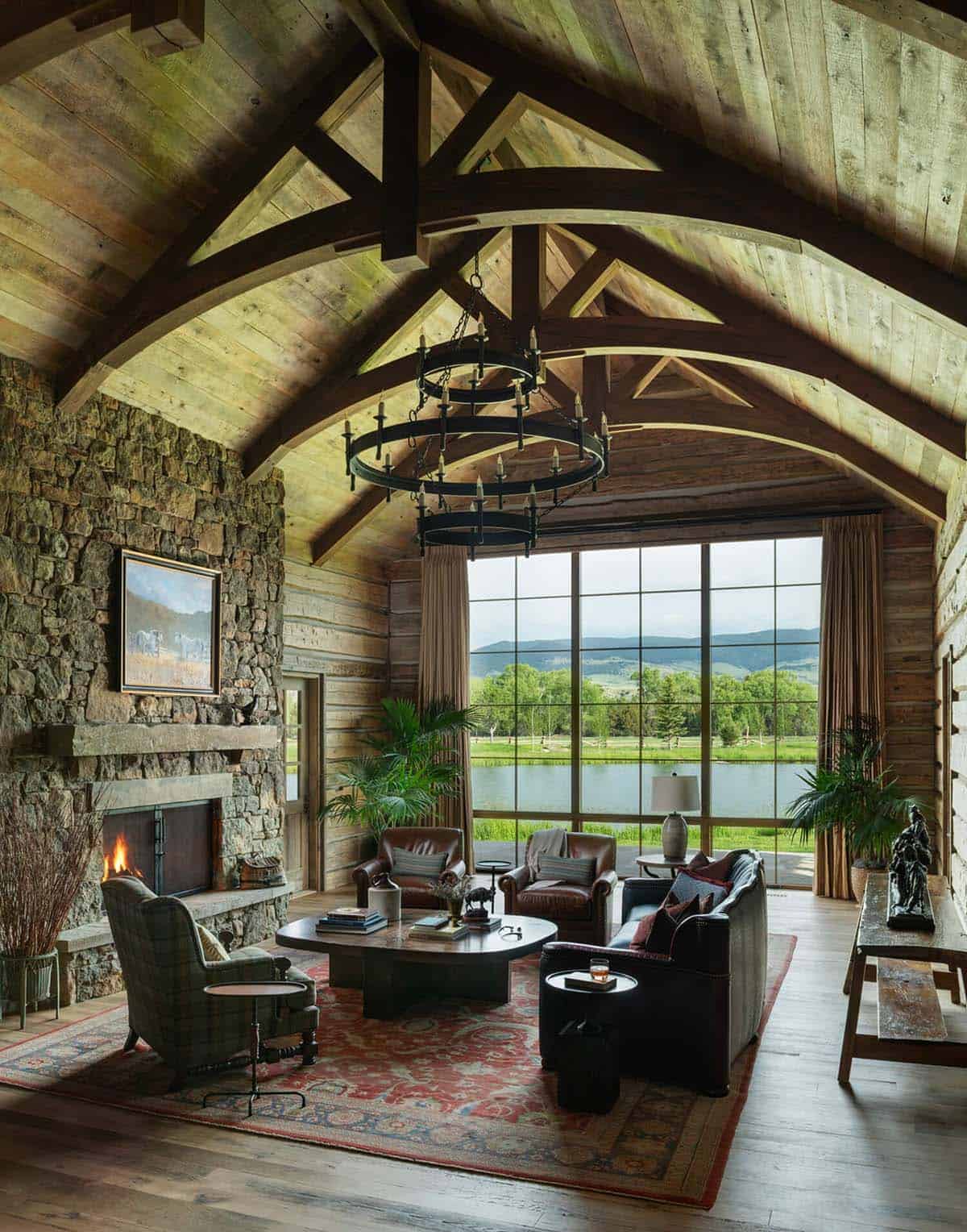 Vaulted log living room with stone fireplace, leather seating, patterned rug, and floor-to-ceiling window over a pond