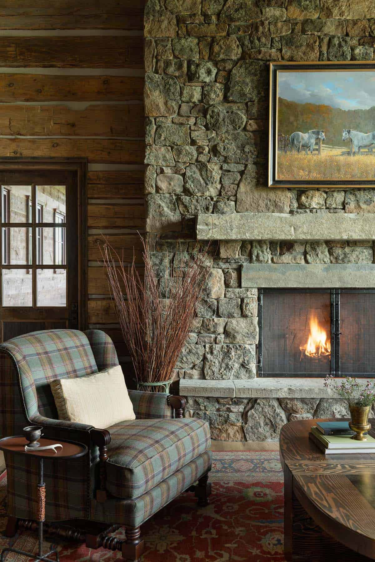 Cozy seating nook with plaid armchair beside a lit stone fireplace and framed horse painting above the mantel