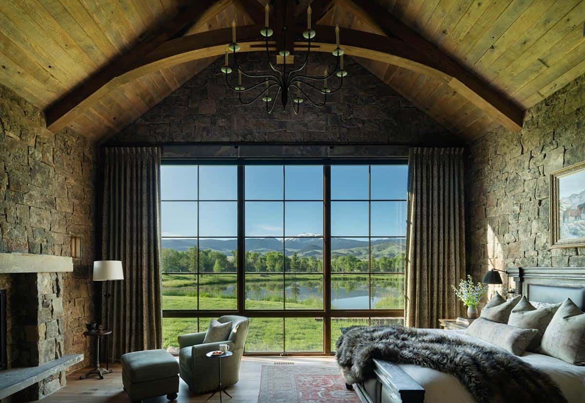 Stone bedroom with vaulted wood ceiling, chandelier, and large window overlooking a pond and mountains