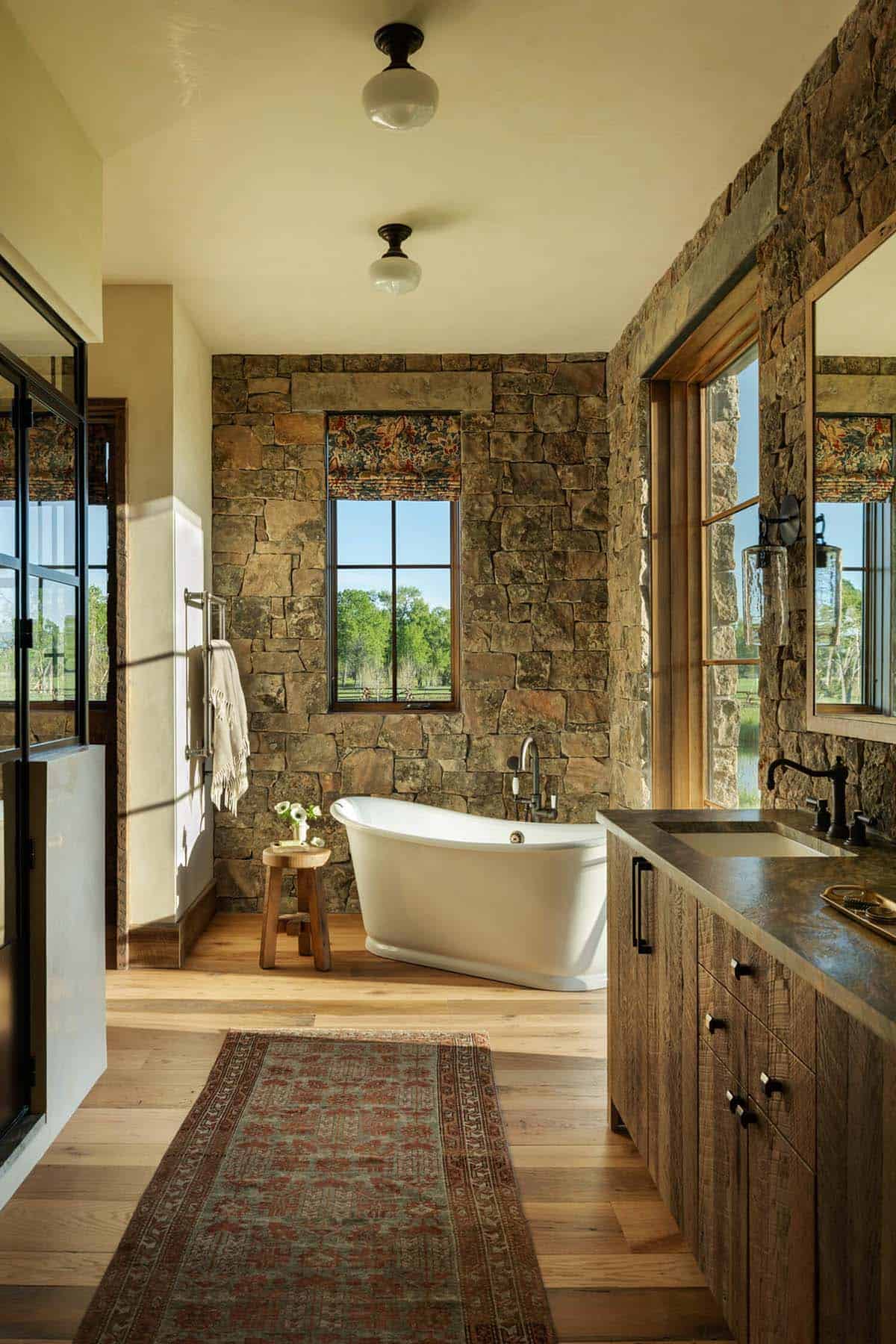 Stone-walled bathroom with freestanding soaking tub, wood vanity, and window views of the ranch grounds
