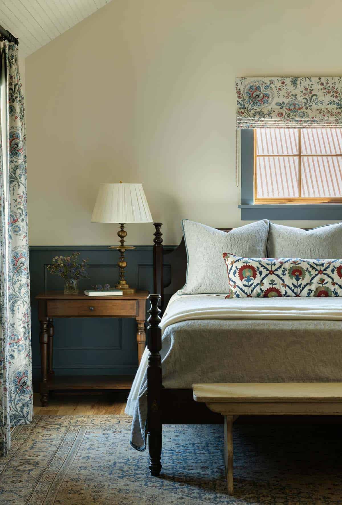 Bedroom nightstand vignette with brass lamp, floral drapery, and blue wainscoting beside a four-poster bed