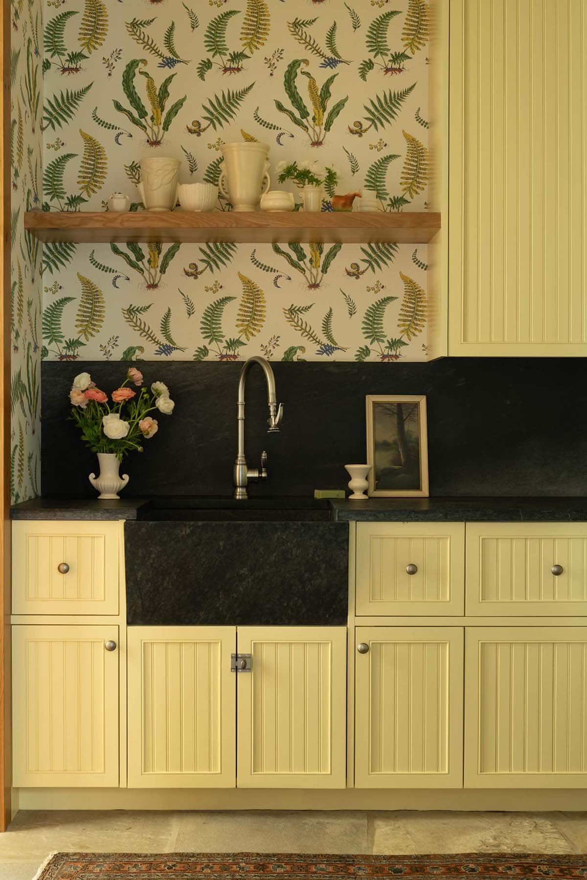 Butler's pantry sink area with fern-print wallpaper, dark soapstone counter, and pale yellow beadboard cabinets