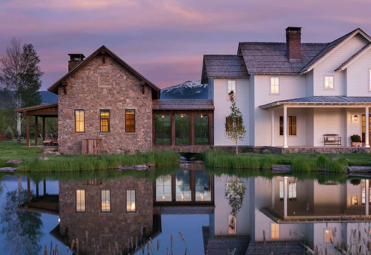 Twilight view of stone and white farmhouse buildings connected by glass breezeway, reflected in a pond
