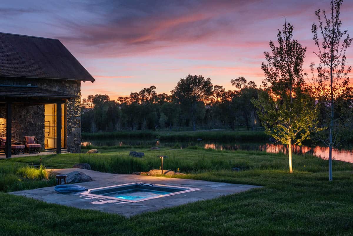 Sunken hot tub glowing at dusk beside a stone guesthouse overlooking a pond and pink sunset sky