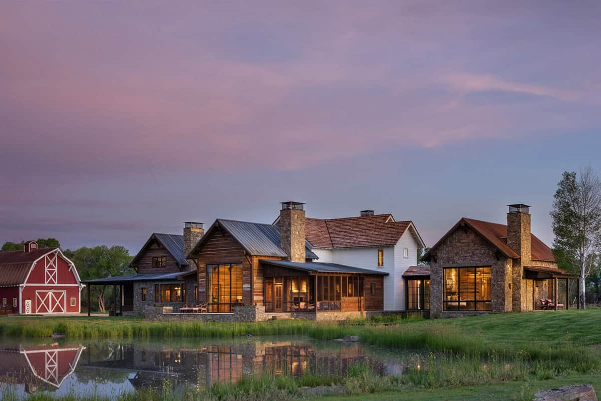 Ranch compound at sunset with red barn, stone-and-wood buildings, and glowing windows reflected in a pond