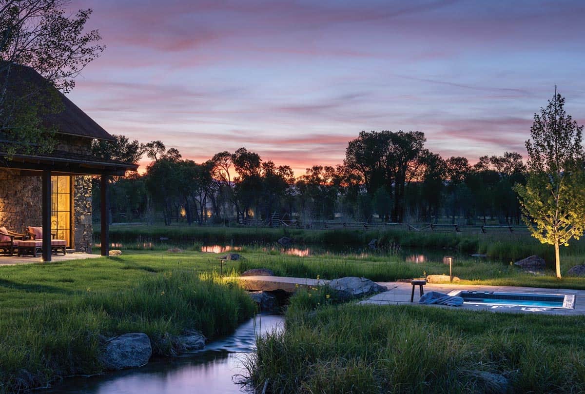 Twilight garden path with stream, stepping stones, and lit hot tub leading toward a stone guesthouse