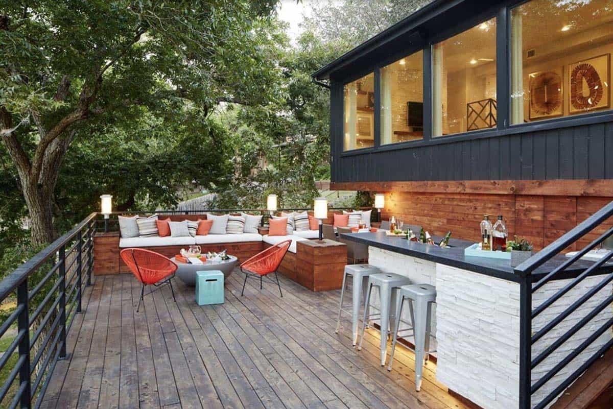 Elevated wood deck with a white stacked stone outdoor bar topped with metal stools, and an L-shaped white sectional with orange and gray pillows, surrounded by tall trees