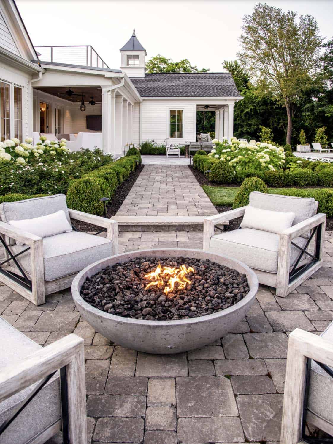 Four gray upholstered lounge chairs with wood frames arranged around a round concrete gas fire pit on a stone patio, with a white farmhouse-style home and manicured gardens in the background