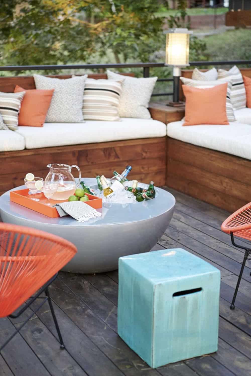 Round concrete coffee table used as a drink station with a pitcher, cocktails, and iced bottles, flanked by orange woven chairs and a turquoise garden stool