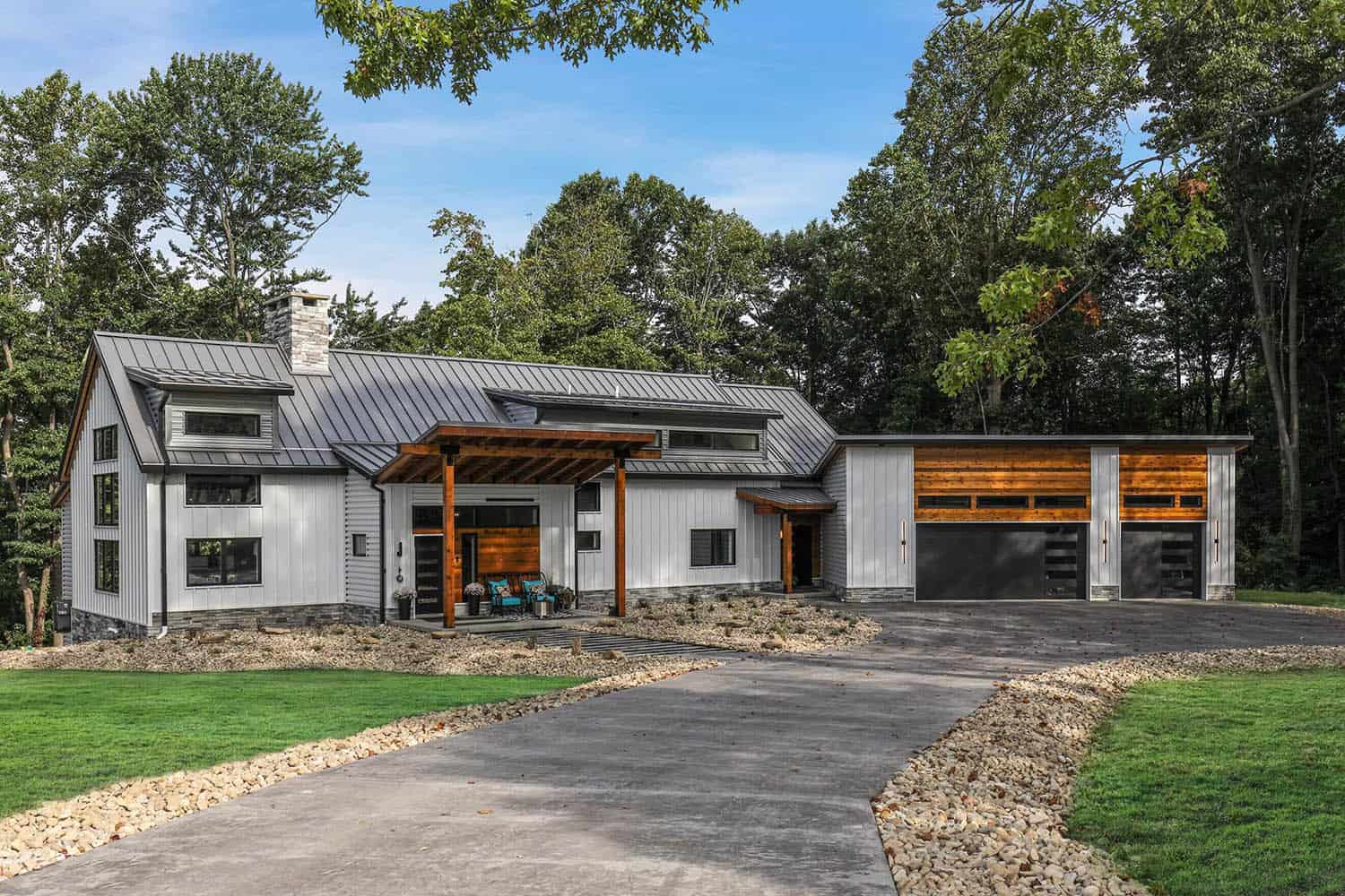 Rustic modern farmhouse exterior with metal roof, board-and-batten siding, wood pergola entry, and attached garage with cedar accents, surrounded by trees