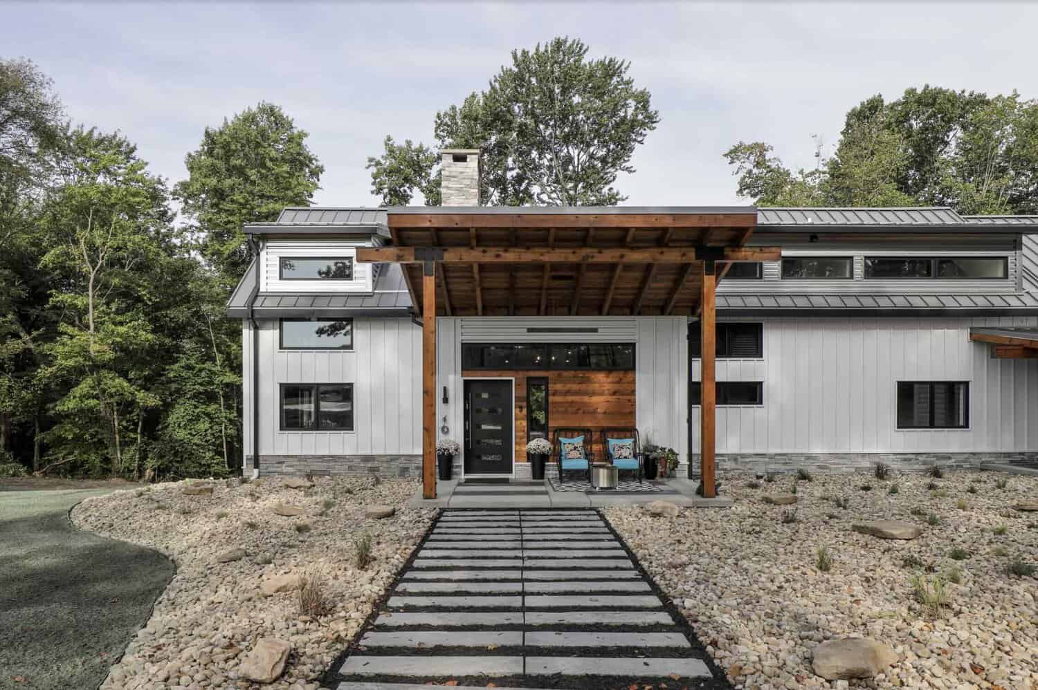 Front entrance with cantilevered wood pergola, black door, cedar siding accent, stone chimney, and linear stepping-stone walkway through river rock landscaping