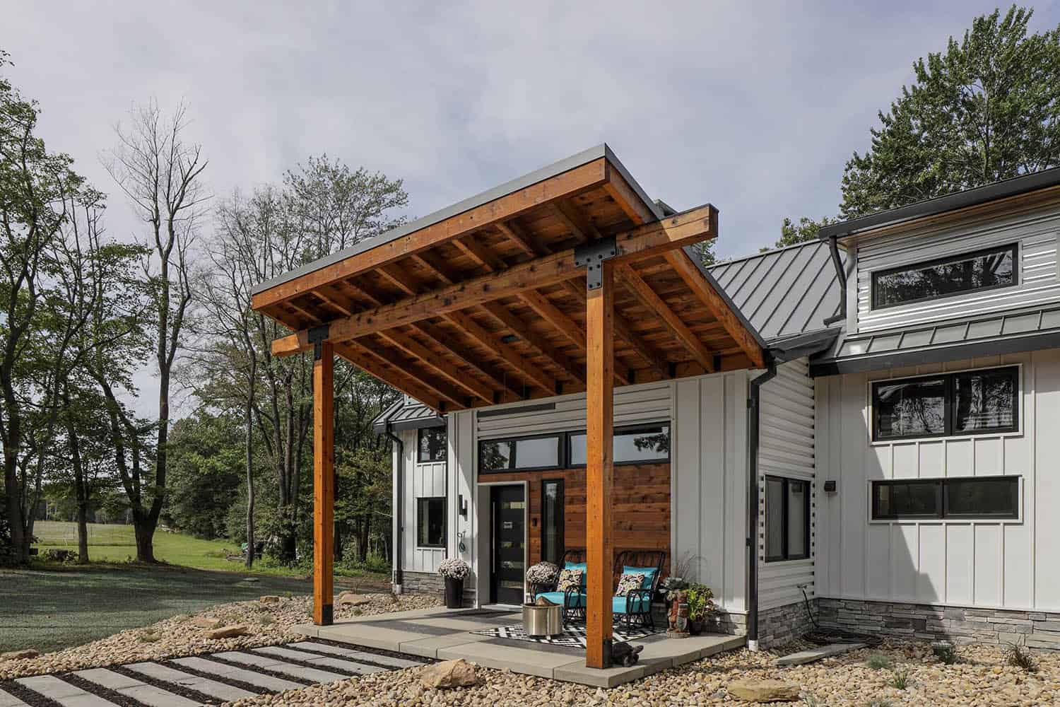Angled view of timber-frame entry pergola with exposed rafters, black steel connectors, and covered porch seating area