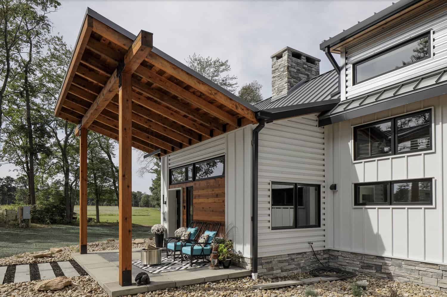 Exterior corner view showing timber pergola, corrugated metal siding, stone chimney, and covered patio with teal outdoor chairs