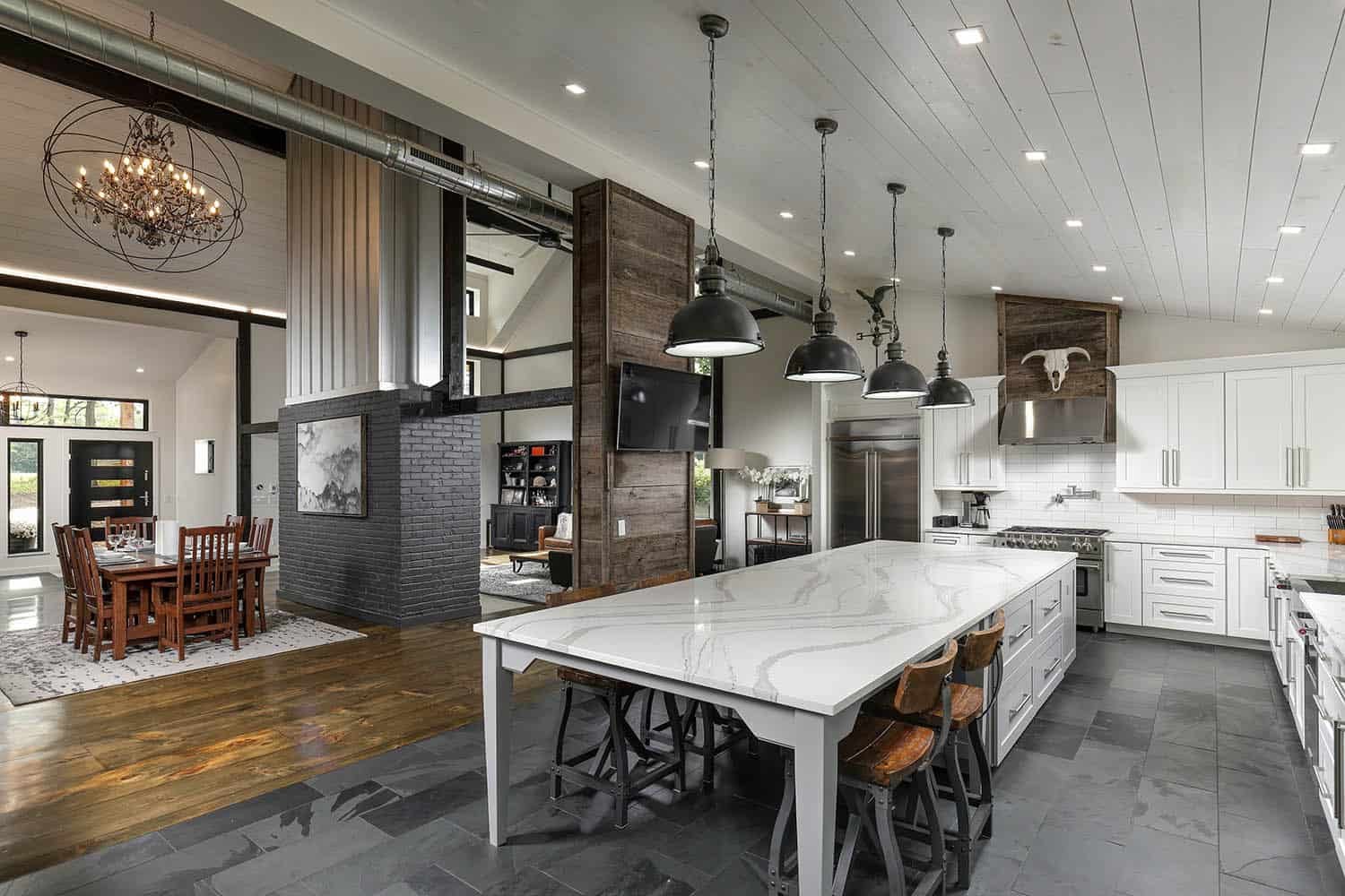 Open-concept kitchen and dining area with white island, industrial pendant lights, exposed ductwork, and dark brick chimney column