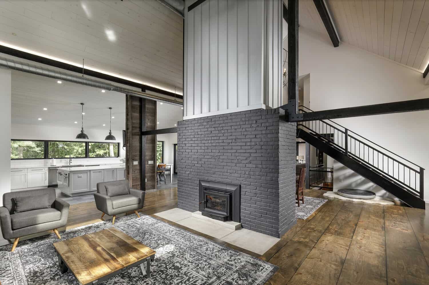 Two-story great room with black-painted brick fireplace column, steel staircase, reclaimed wood floors, and gray armchairs