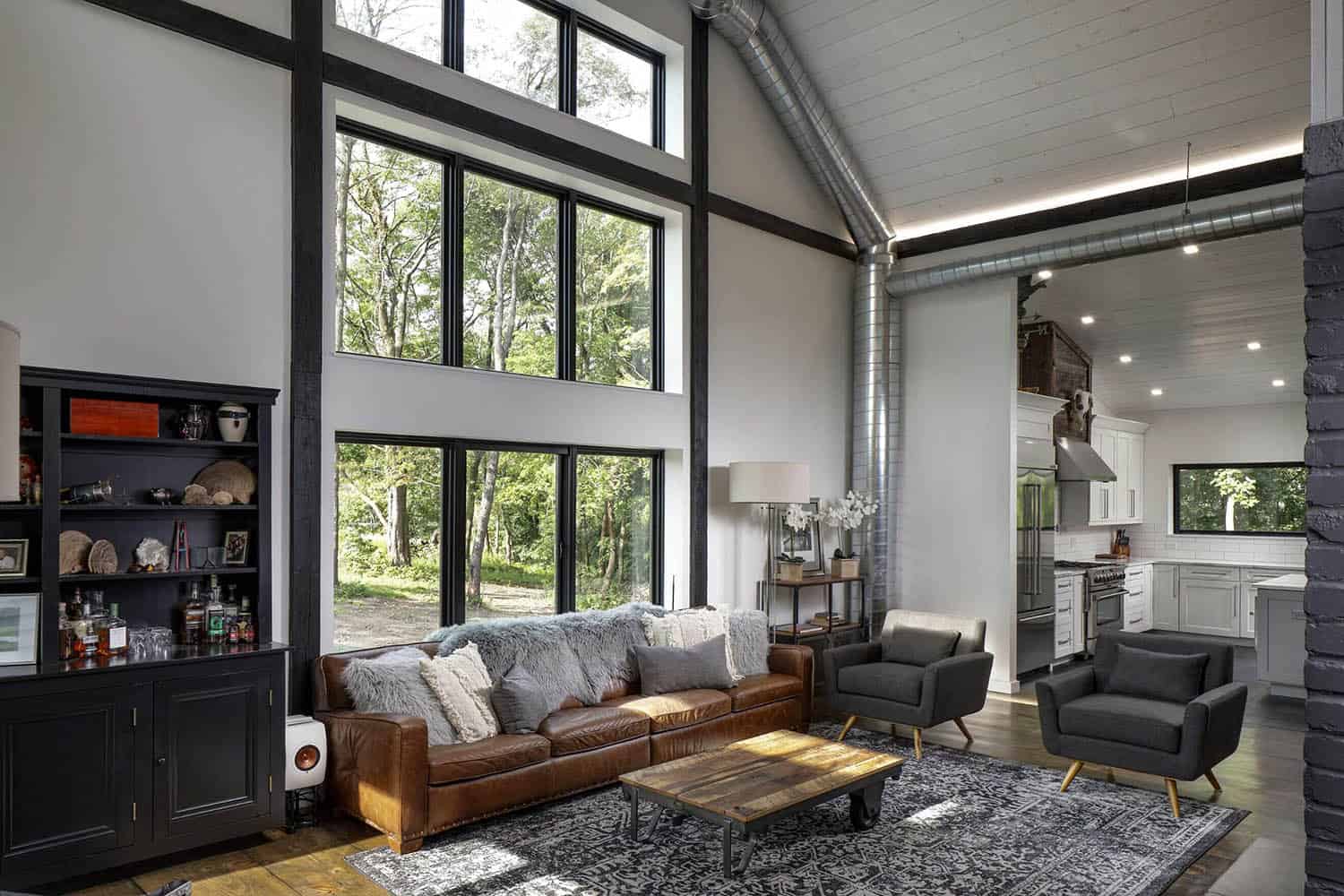 Living room with tall black-framed windows, leather sectional sofa, black brick column, and view into white kitchen