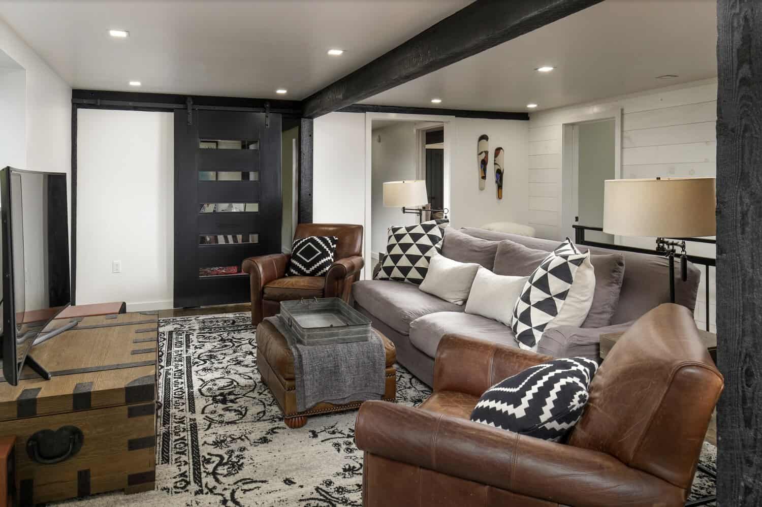 Cozy den with gray sectional sofa, leather armchairs, patterned throw pillows, black sliding barn door, and wood trunk coffee table