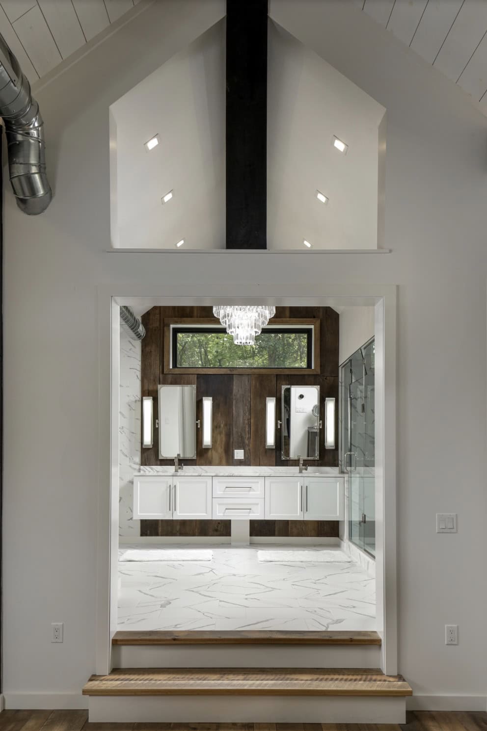 View through a gabled interior window frame into a bathroom with wood-paneled vanity wall and crystal chandelier
