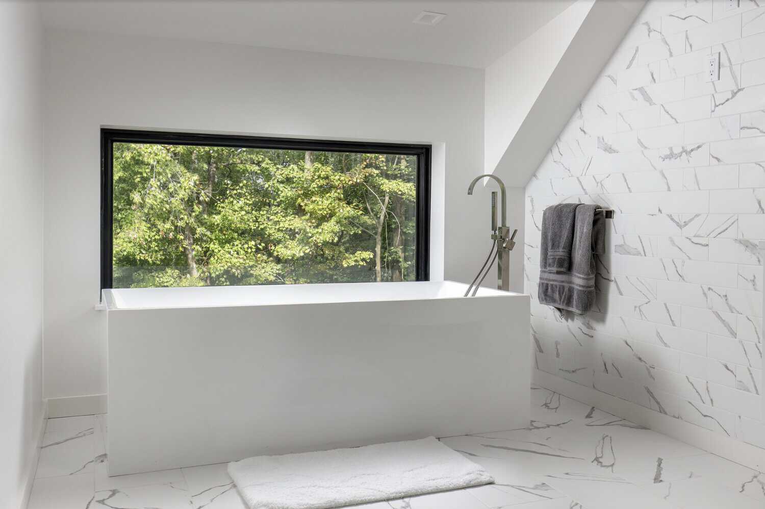Minimalist bathroom with rectangular soaking tub under a large window, marble subway tile wall, and gray towels