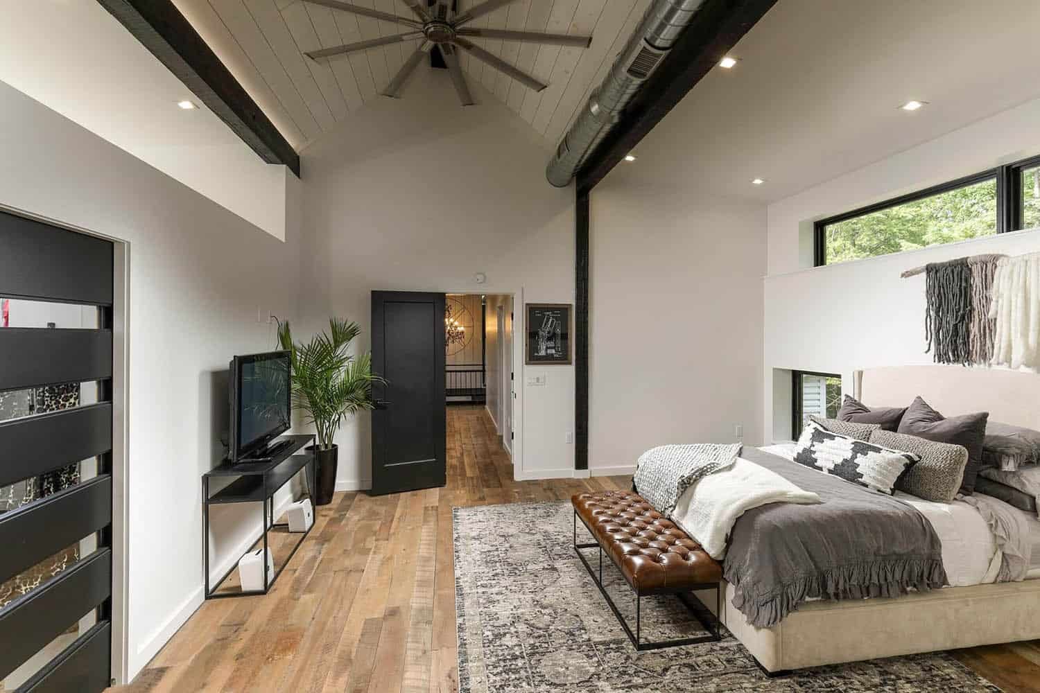 Primary bedroom with vaulted wood ceiling, black beams, ceiling fan, leather bench, and hallway view to staircase