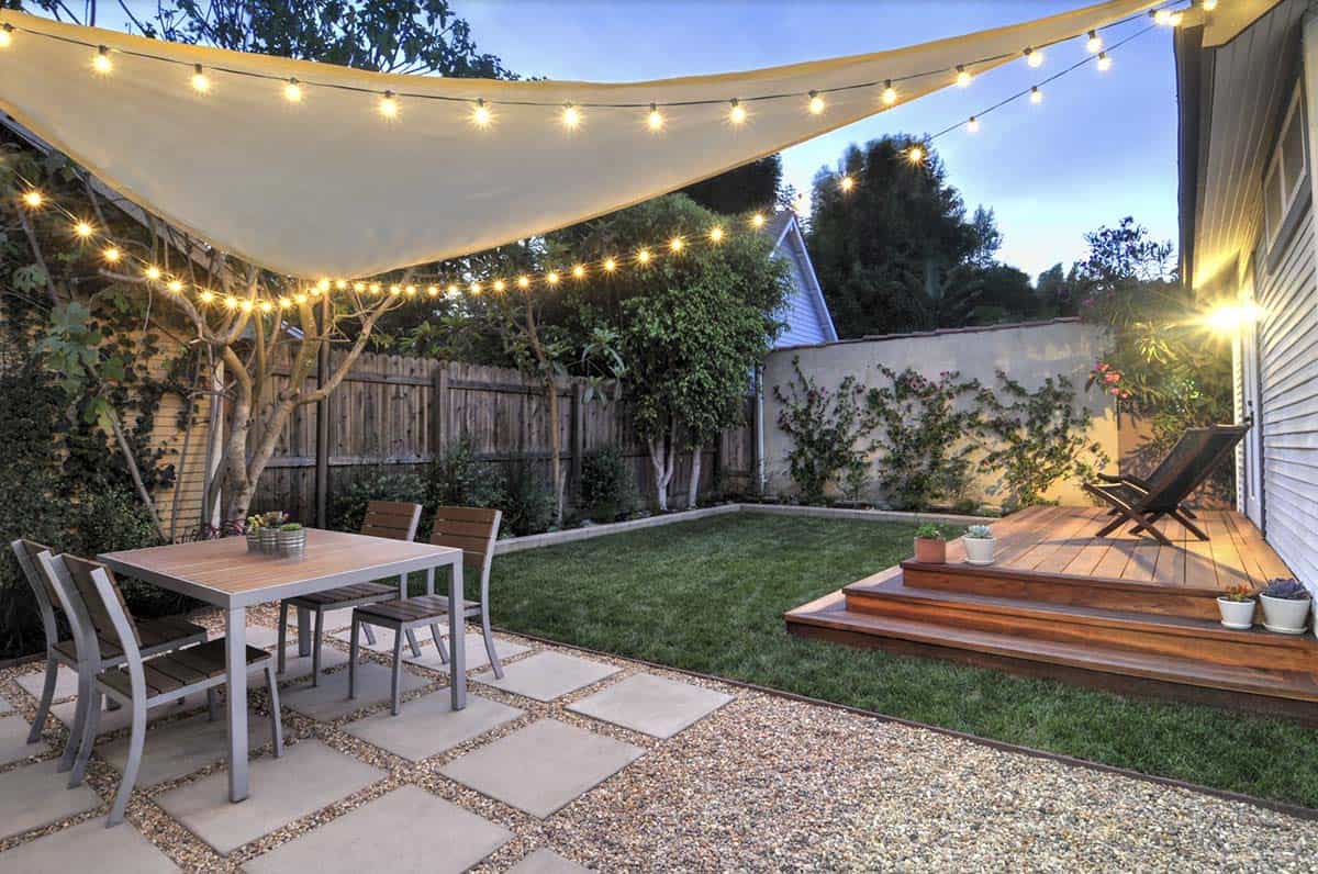 Backyard dining table set on paver and gravel patio, shaded by a cream fabric sail draped with string lights, with a lawn and wood deck visible beyond