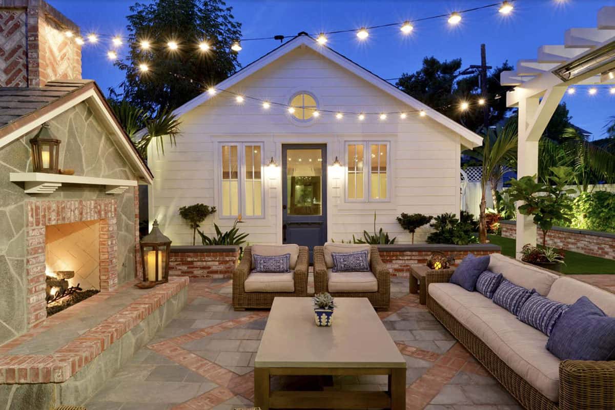 Evening patio with a brick and stone outdoor fireplace, wicker sofa and chairs with blue pillows, a patterned paver floor, and string lights strung across a white cottage facade