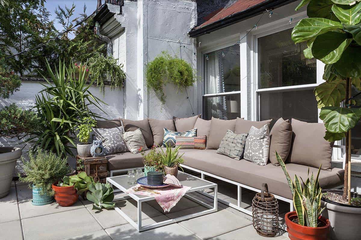 Small patio with taupe L-shaped sectional, patterned throw pillows, glass coffee table, string lights, and potted plants including a fiddle leaf fig and snake plants