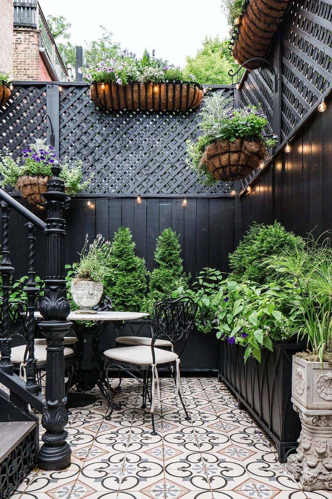 Narrow row house patio with black lattice fencing, hanging coco-lined flower baskets, string lights, potted topiaries, a marble bistro table with mismatched black chairs, and patterned cement tile floors