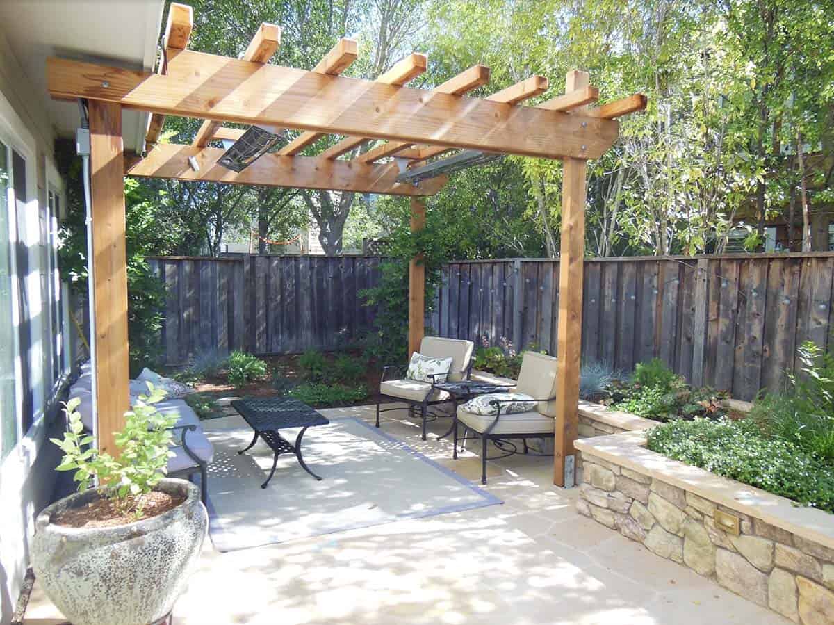 Small backyard patio with a wooden pergola, black wrought-iron seating with cushions, flagstone paving, and raised stone-veneered planting beds along a wood fence
