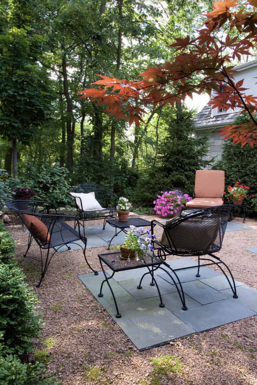 Shaded garden patio with black wrought-iron chairs and side tables on scattered flagstone pavers set into gravel, surrounded by lush trees, a Japanese maple, and potted flowers
