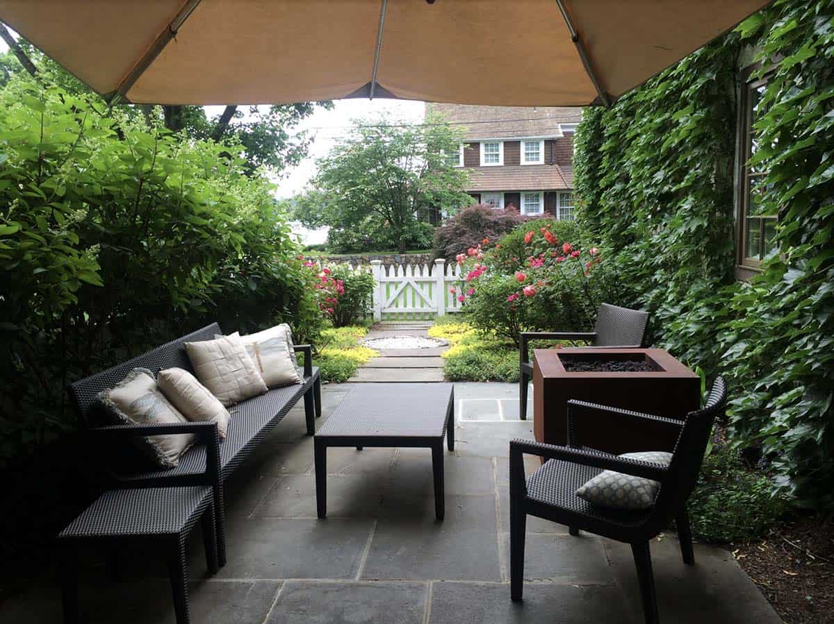 Small flagstone patio with black wicker sofa and chairs, throw pillows, a square copper fire pit, and a large umbrella, framed by ivy-covered walls and a garden gate