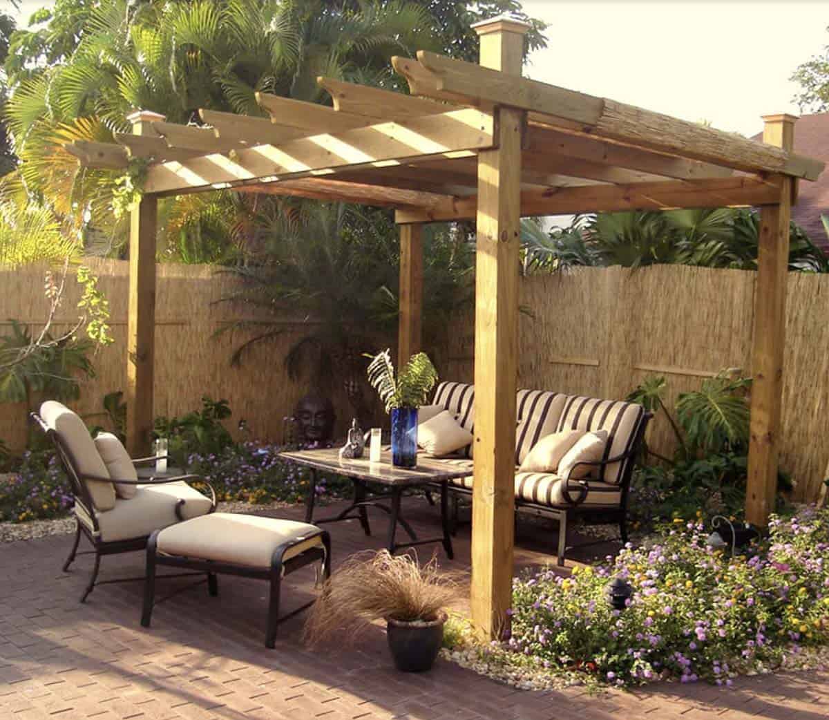 Small brick paver patio with a natural wood pergola, black wrought-iron sofa and chairs with striped cushions, a glass-top coffee table, and a garden bed of purple flowers