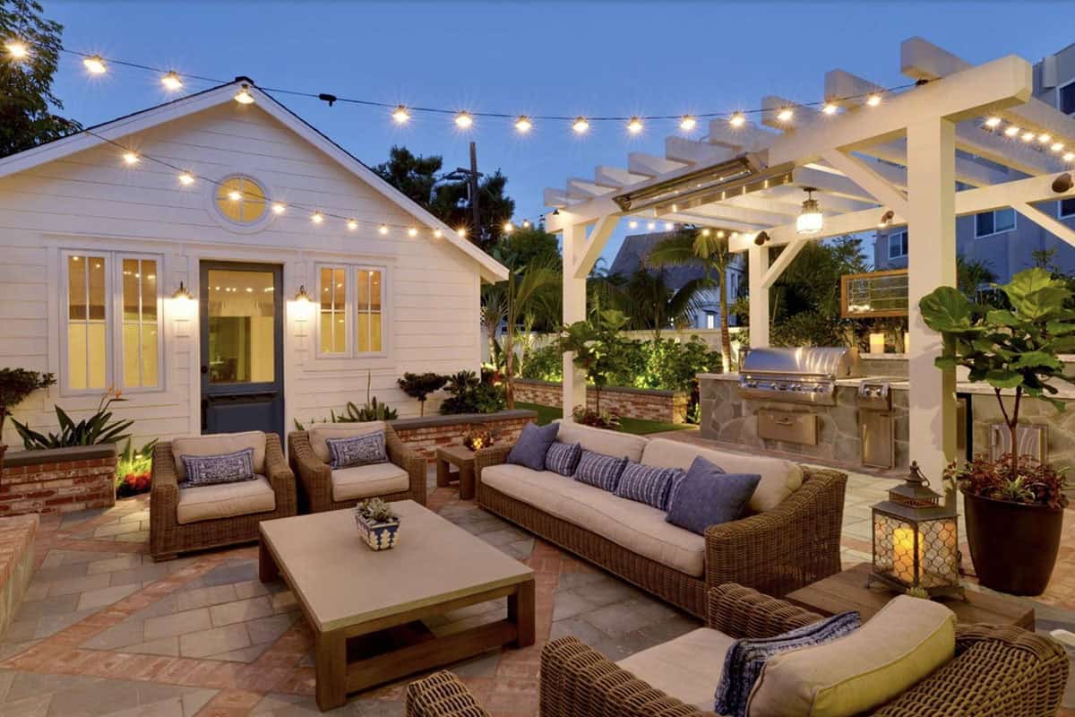 Twilight patio with woven wicker sofa and chairs, blue and white pillows, a wooden coffee table, string lights overhead, a white pergola with an outdoor kitchen, and a stone-and-brick fireplace