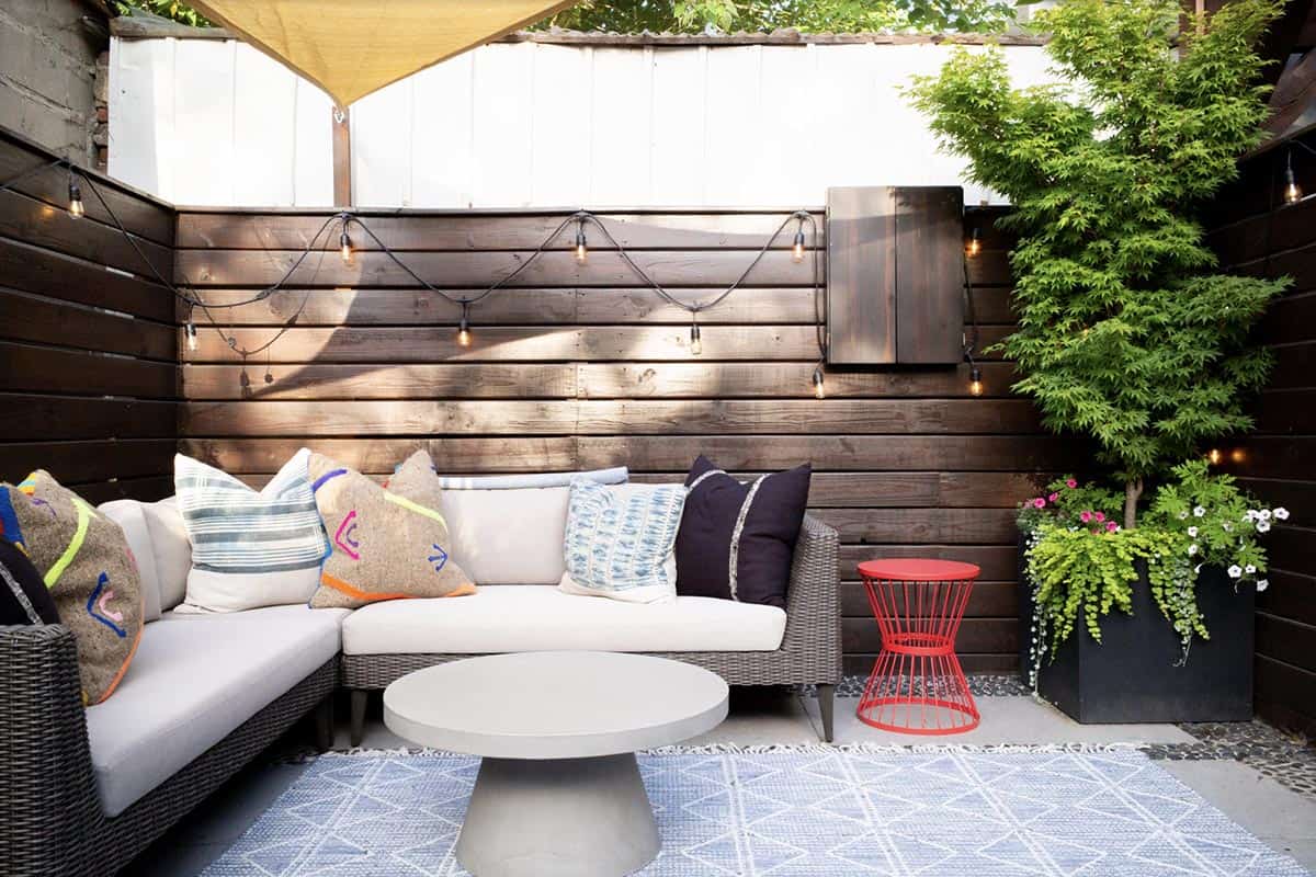 Small backyard patio with a dark stained wood fence, white sectional sofa with colorful throw pillows, round concrete coffee table, red metal side table, blue patterned rug, and a Japanese maple in a black planter