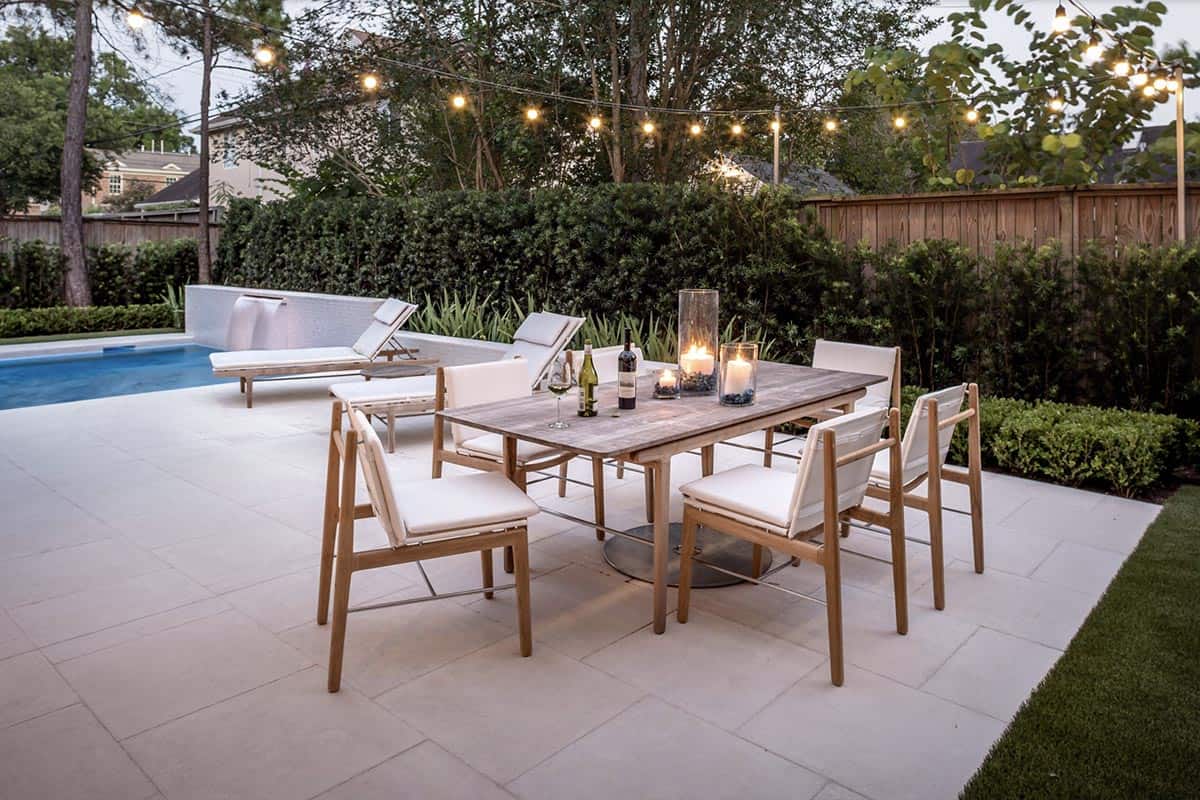 Poolside patio with a white travertine floor, wooden dining table and chairs with white cushions, lit candles, string lights, and lounge chairs beside a pool with a water feature