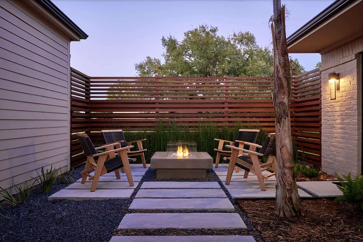 Modern side yard patio with concrete pavers set in dark gravel, a square concrete fire pit with a glass wind guard, teak and wicker armchairs, and a horizontal wood slat privacy fence