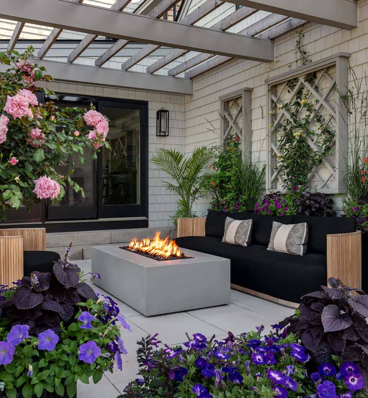 Modern farmhouse patio with a black sofa and patterned pillows, a concrete fire pit, trellises with climbing vines, and containers of purple petunias and pink roses under a pergola