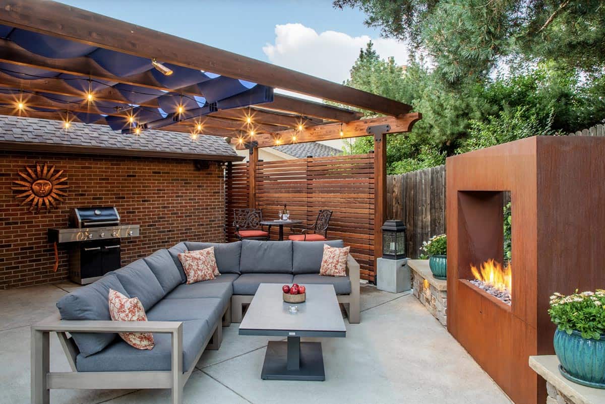 Patio with a gray sectional sofa, brick outdoor kitchen with grill, wood pergola with retractable blue wave shades and string lights, slatted privacy screens, and a Cor-Ten steel outdoor fireplace
