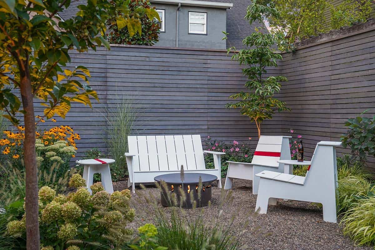 Small pea gravel patio with white modern Adirondack-style chairs, a black round fire pit, gray wood privacy fence, and garden plantings including black-eyed Susans and ornamental grasses
