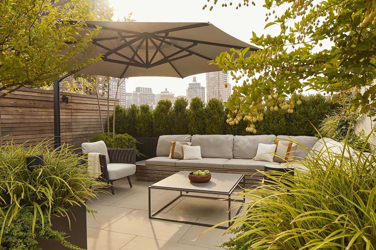 Rooftop patio with a gray sectional sofa and armchair, square metal-framed coffee table, large cantilever umbrella, wood privacy fence, and ornamental grasses and hedges, with a city skyline in the background