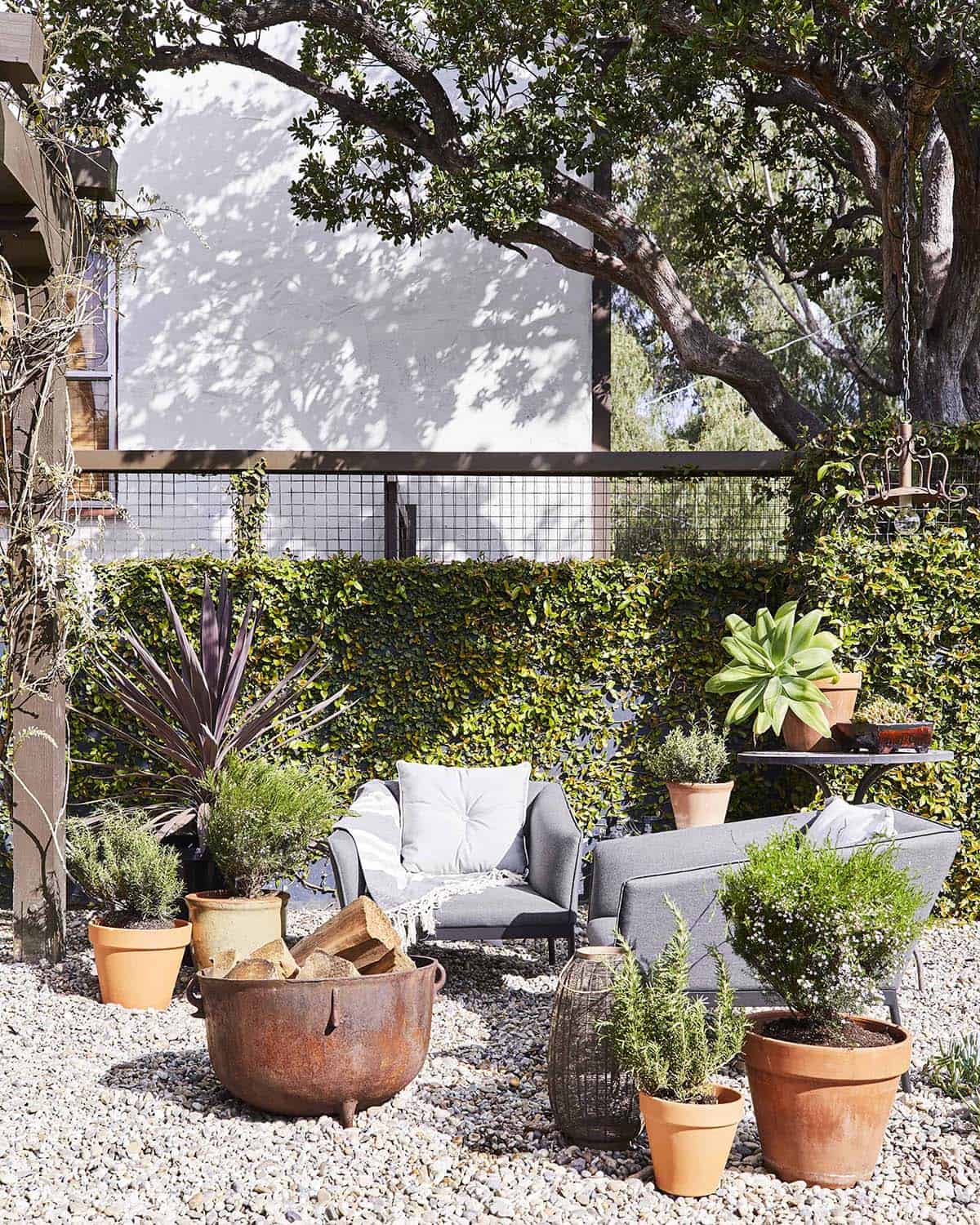 Small gravel patio shaded by a tree, with gray Article lounge chairs, a rusted iron cauldron used as a DIY fire pit, terracotta planters of varying sizes, and a vintage iron pendant light hanging from a branch