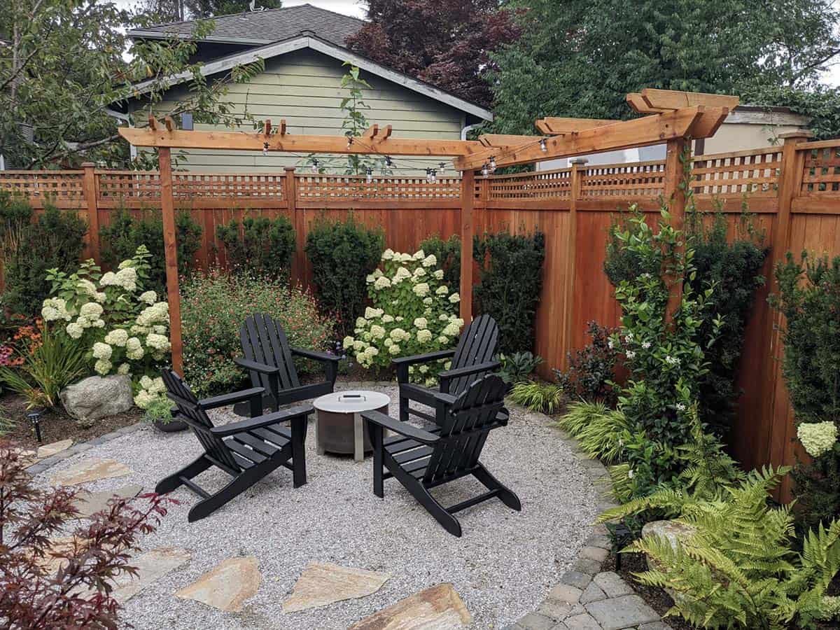 Circular gravel firepit patio with black Adirondack chairs, a round metal fire pit, stone pavers, a cedar arbor with string lights, and a tall cedar privacy fence surrounded by hydrangeas and ferns