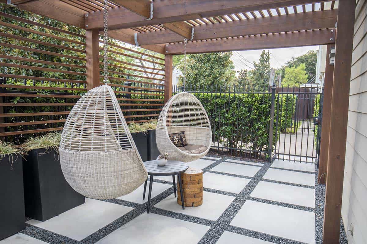Covered patio with two white wicker hanging egg chairs, a small round side table, concrete pavers set in dark gravel, a wood slat pergola and privacy screen, and a black iron gate