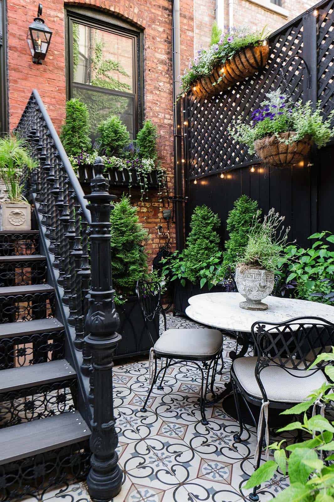 Small brownstone patio with patterned cement tile flooring, black wrought-iron bistro table and chairs, a black lattice privacy fence with hanging flower baskets, and a black iron staircase