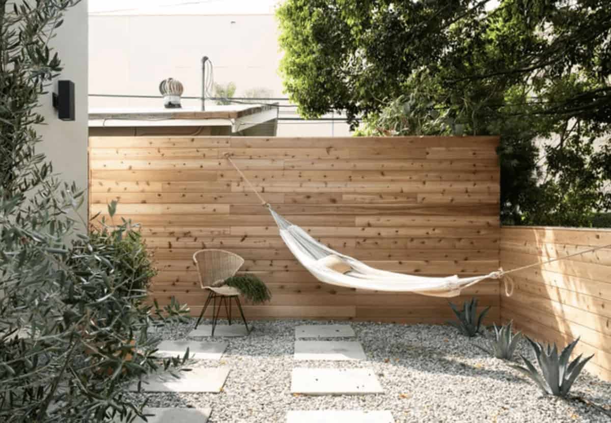 Small gravel patio with a white hammock hung against a wood plank fence, a woven wicker chair, concrete pavers, agave plants, and an olive tree.