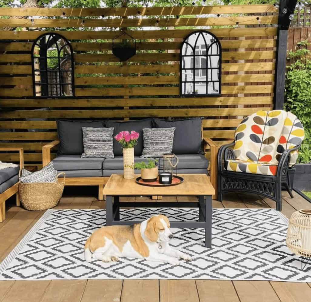 Wood deck patio with a gray sectional sofa, patterned rattan armchair, wood coffee table, black-and-white geometric rug, arched mirrors on a wood slat wall, and a beagle lounging on the rug