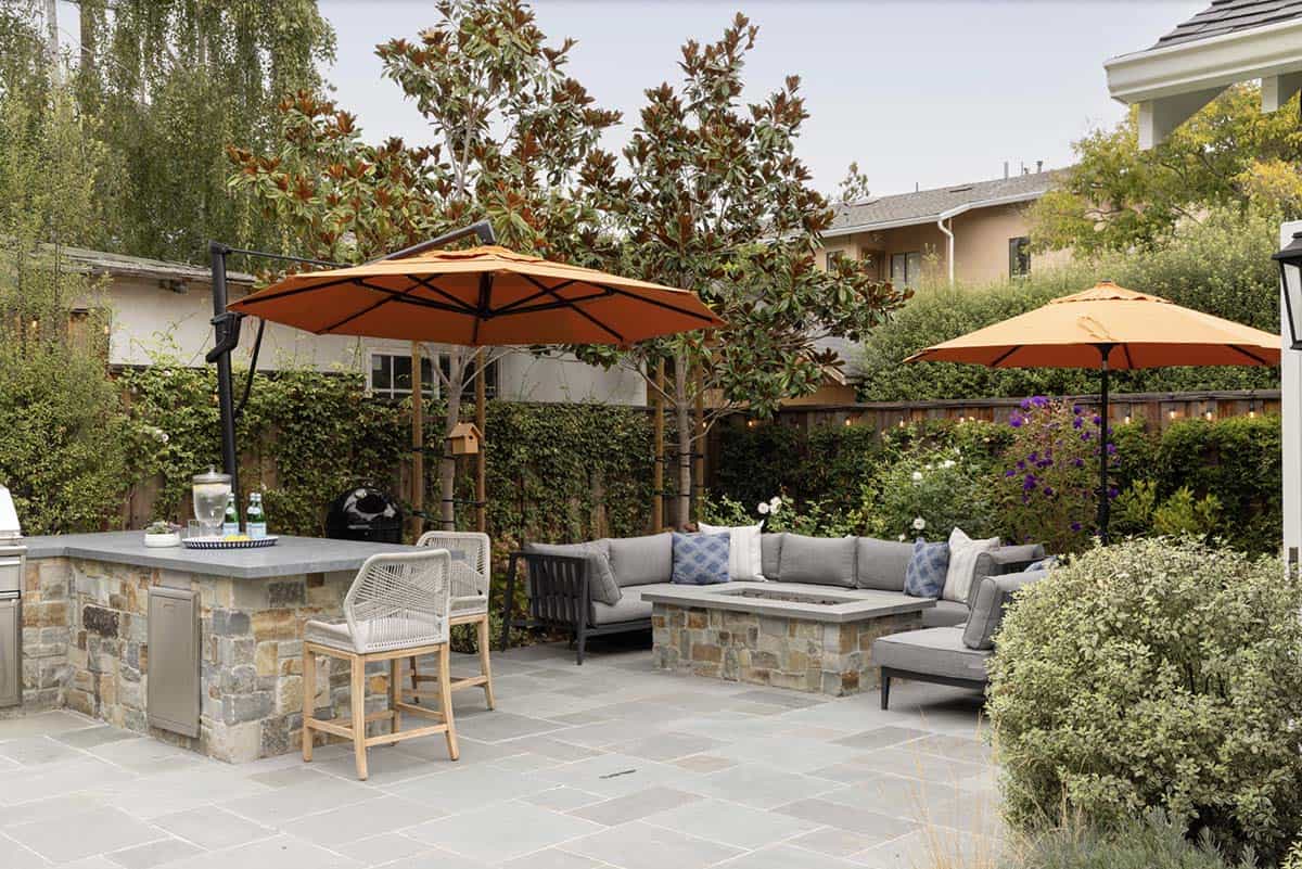 Stone patio with a built-in stone fire pit surrounded by a gray sectional sofa, two burnt orange cantilever umbrellas providing shade, and a stone bar counter with wicker stools in the foreground