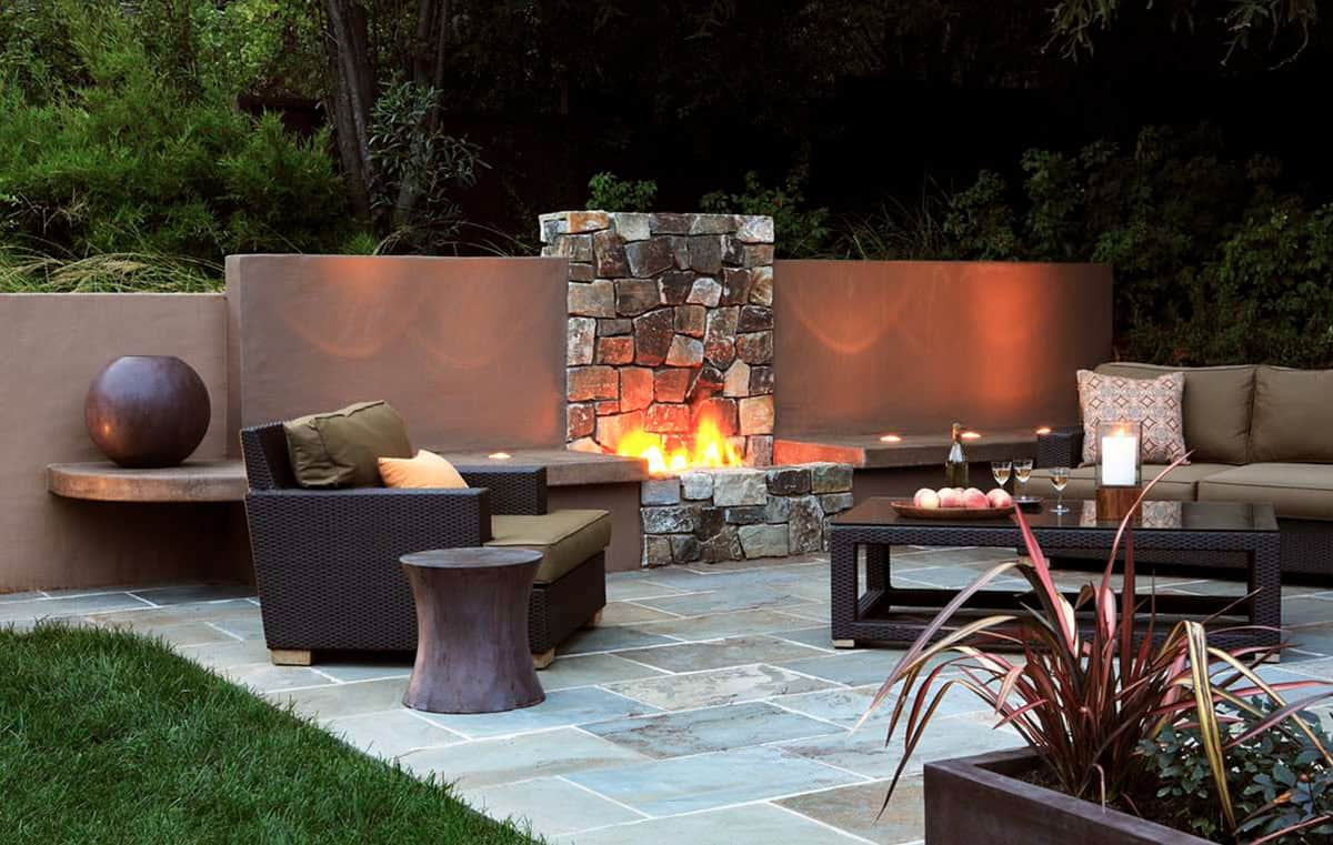 Evening patio with a tall stone outdoor fireplace flanked by curved stucco walls, dark wicker lounge chairs and a sectional, a low coffee table set with wine and fruit, and candlelight throughout