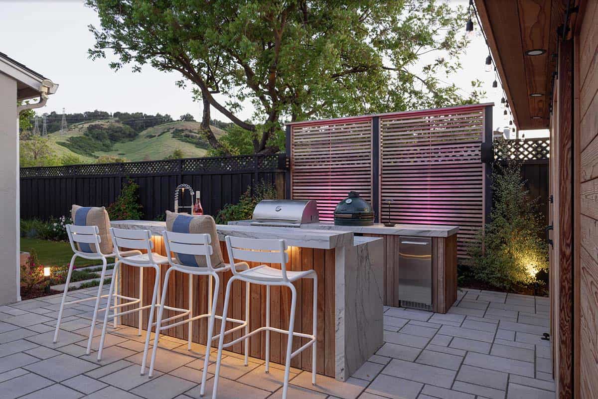 Outdoor kitchen with a stone waterfall-edge island, white bar stools, a built-in grill and ceramic smoker, and a wood slat privacy screen backlit with pink lighting